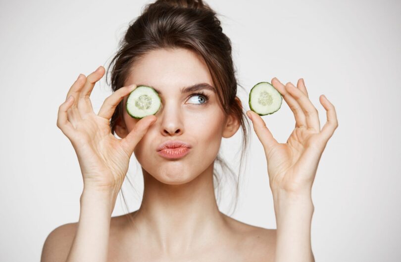 Young beautiful naked girl smiling hiding eye behind cucumber slice over white background. Beauty spa and cosmetology concept. Copy space.